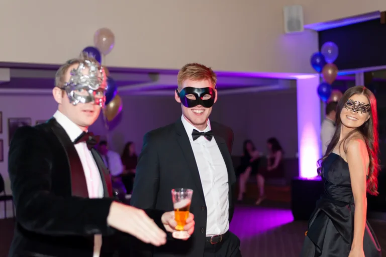 kooyong lawn tennis club masquerade ball candid three guests posing Three guests pose together at the YarraYong Ball at Kooyong Lawn Tennis Club in Toorak, smiling broadly and enjoying the festive evening. The man in the center wears a black tuxedo with a black butterfly mask and holds a glass of red beverage, the man on the left wears a black velvet tuxedo with a silver mask, and the woman on the right wears a black dress with a lace mask. Warm string lights and purple uplighting create a sophisticated masquerade ball atmosphere.