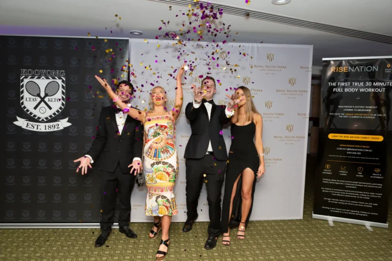 kooyong lawn tennis club masquerade ball celebration with confetti Four guests celebrate energetically at the Kooyong Lawn Tennis Club End-of-Year Masquerade Ball in Toorak. Two men in black tuxedos with bow ties and face paint gesture toward the camera with wide smiles, while a woman in a colorful fan-print dress raises her hands in cheer, and another woman in a black dress with a thigh-high slit holds a drink. Purple and gold confetti falls around them against a backdrop of Kooyong and Royal South Yarra banners, capturing a festive and glamorous party atmosphere.