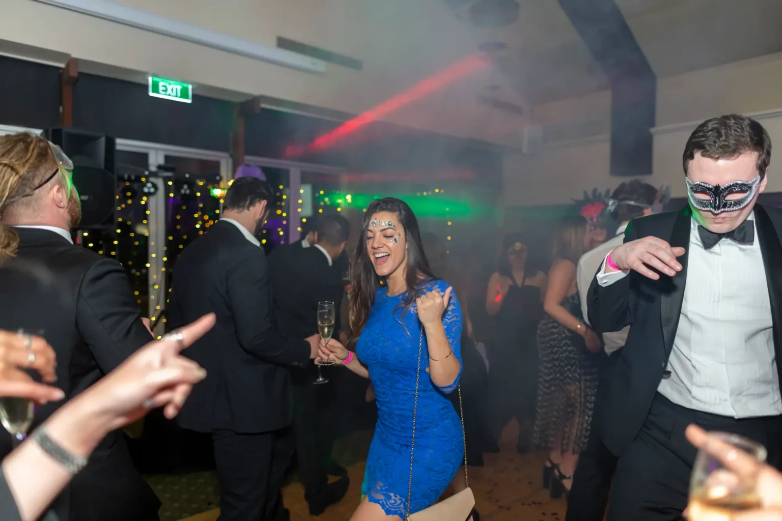 Guests dancing energetically on the dance floor at Kooyong Lawn Tennis Club Masquerade Ball, with vibrant red and green stage lights, smoke effects, and a woman in a blue lace dress laughing and raising her fist, while men in tuxedos and masquerade masks join the celebration