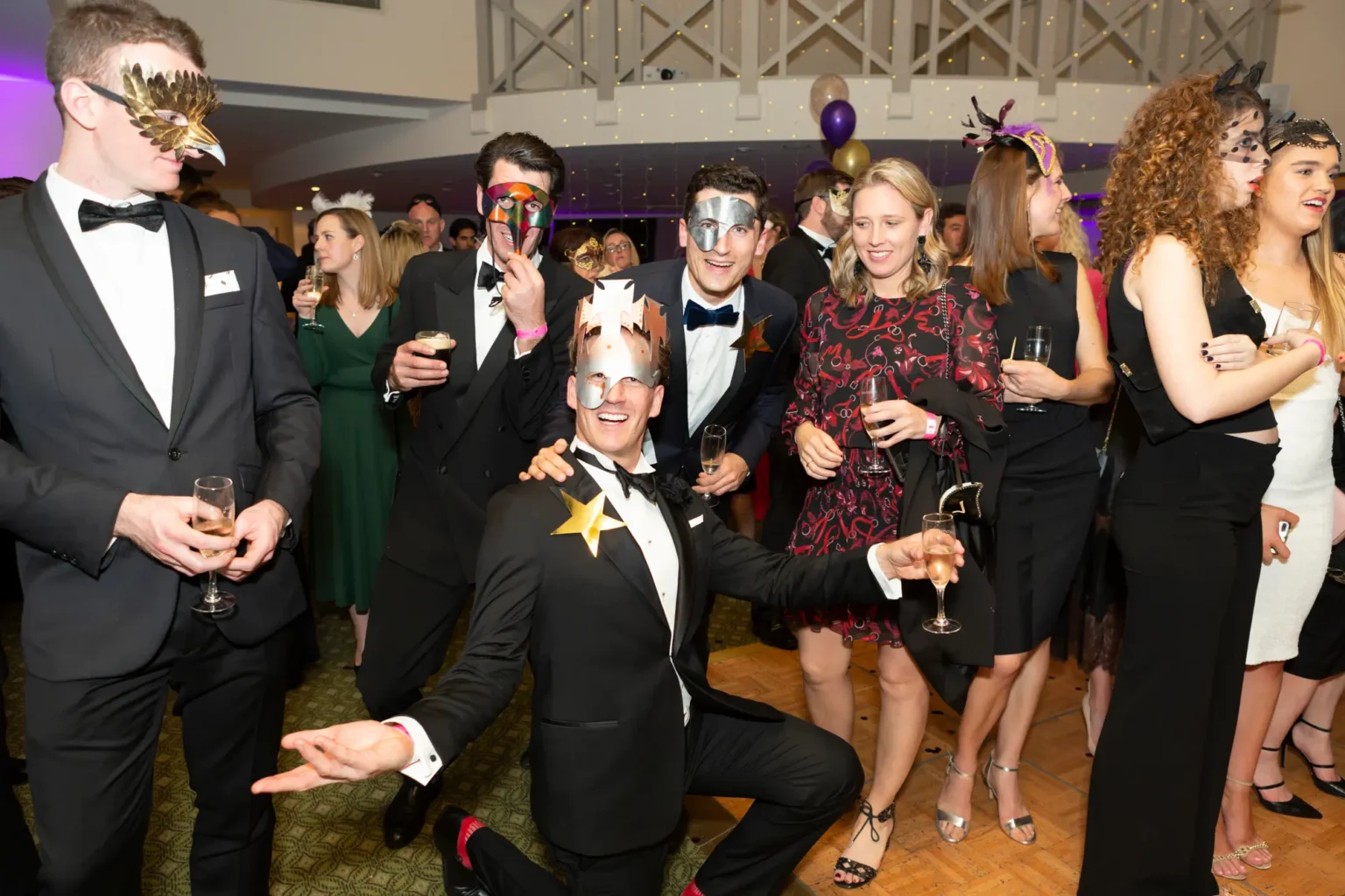 A lively group poses at the Kooyong Lawn Tennis Club End-of-Year Masquerade Ball in Toorak. Three men in tuxedos and elaborate masquerade masks, including a crouching man with a wide smile, are in the foreground, while women in elegant dresses and masks stand behind them holding glasses. The indoor venue features fairy lights, balloons, and a balcony, creating a festive and joyful atmosphere.