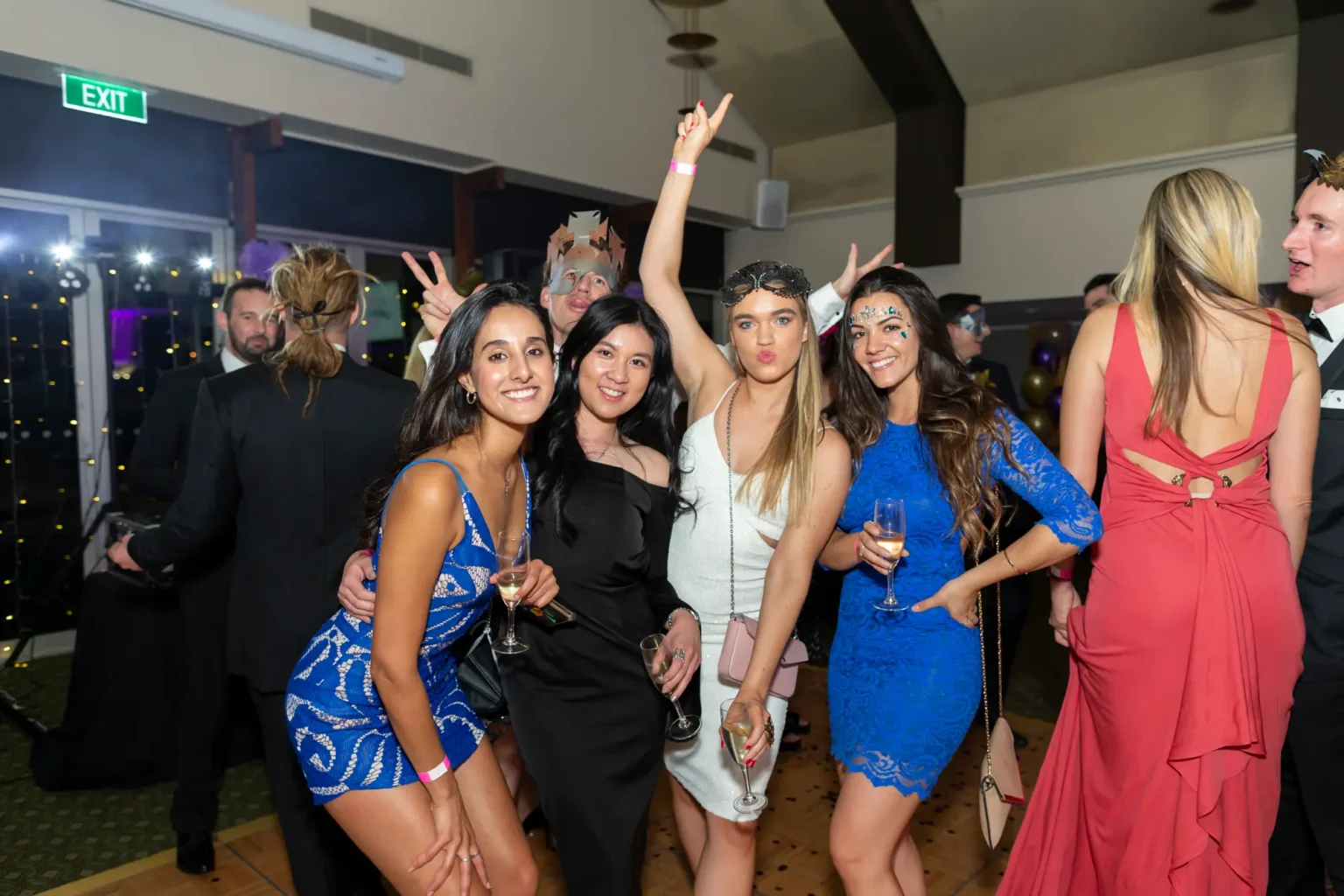 Four women pose energetically during the Kooyong Lawn Tennis Club End-of-Year Masquerade Ball in Toorak, smiling, crouching, and raising a peace sign while holding drinks. Captured in warm evening light with other guests behind them, the image reflects the fun, social atmosphere of the YarraYong Ball celebration.