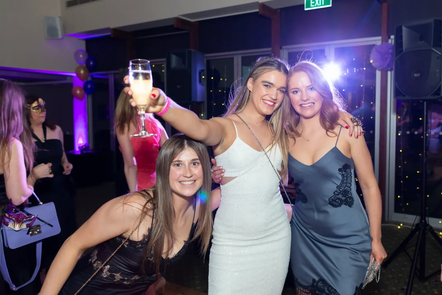 Three women celebrate at the Kooyong Lawn Tennis Club End-of-Year Masquerade Ball in Toorak. One woman in a blue dress crouches low smiling, a woman in black holds a glass, a woman in white raises her arm with a peace sign, and a woman in a blue lace dress poses with her hand on her hip and festive face gems. The lively indoor venue features warm lighting, string lights, and other guests in the background, capturing a joyful and energetic party atmosphere.