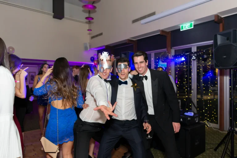 kooyong lawn tennis club masquerade ball energetic mens group party moment Three men in tuxedos and masquerade masks pose energetically at the Kooyong Lawn Tennis Club End-of-Year Masquerade Ball in Toorak, with one crouching forward in a playful gesture. Other guests smile behind them under warm lighting, capturing the lively social atmosphere of the YarraYong Ball celebration.