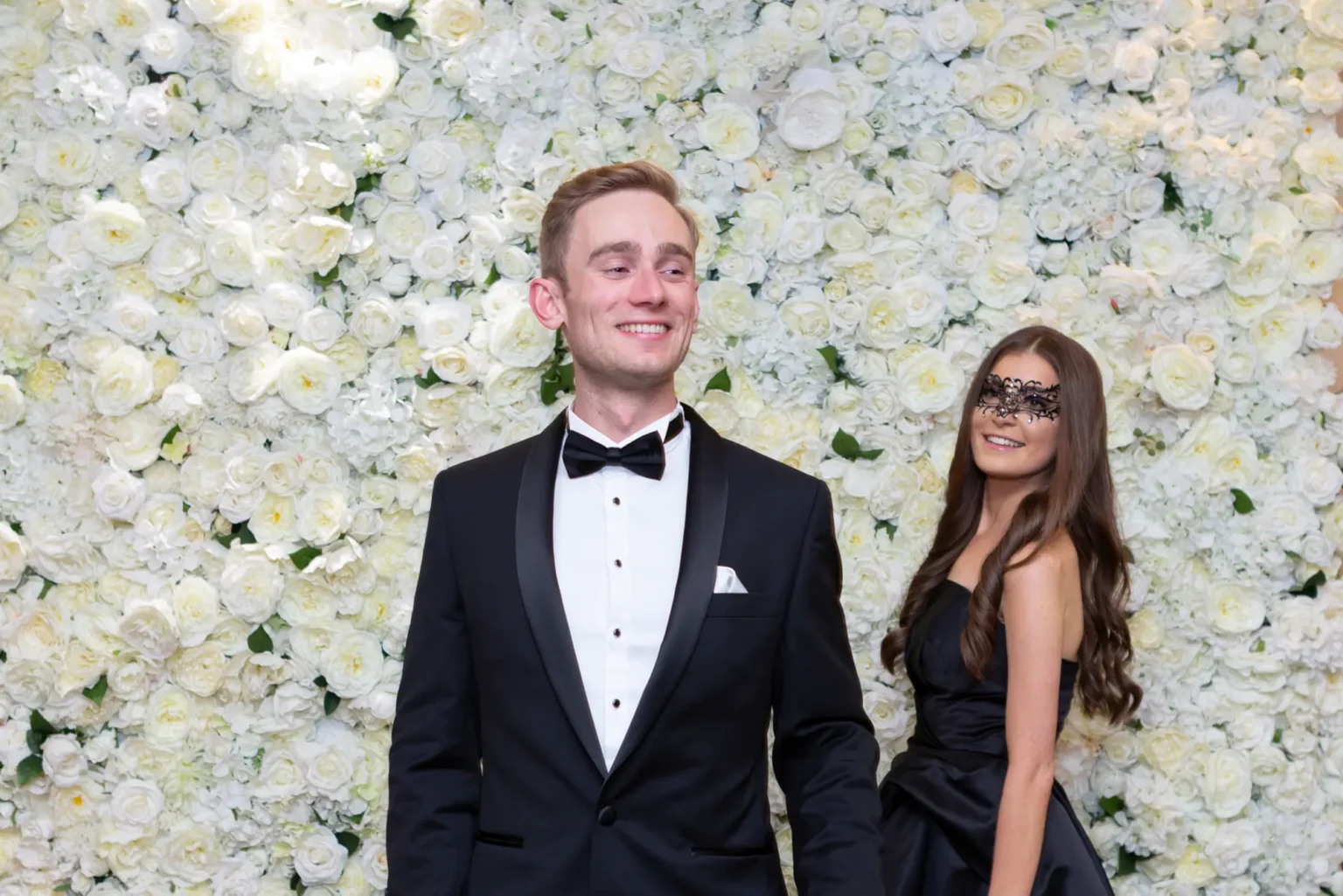 An elegant couple poses in front of a white flower wall at the Kooyong Lawn Tennis Club End-of-Year Masquerade Ball. The man wears a classic black tuxedo and smiles slightly off-camera, while the woman in a strapless black dress and black lace masquerade mask looks up at him warmly. The floor-to-ceiling floral backdrop creates a bright, romantic contrast to their dark formal attire, capturing a joyful and sophisticated moment from the glamorous celebration.