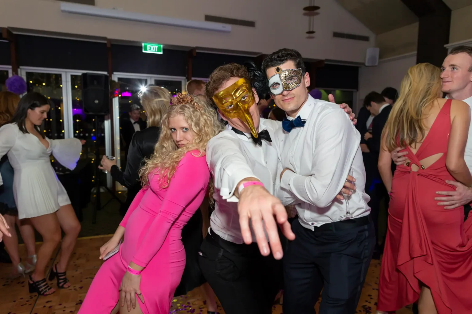 Three guests pose energetically at the Kooyong Lawn Tennis Club End-of-Year Masquerade Ball, with a man in a gold bird-beak mask pointing toward the camera between a woman in a bright pink dress and a man in a silver mask. Dark wood paneling, speakers, and mingling guests in the background highlight the lively black-tie party atmosphere.