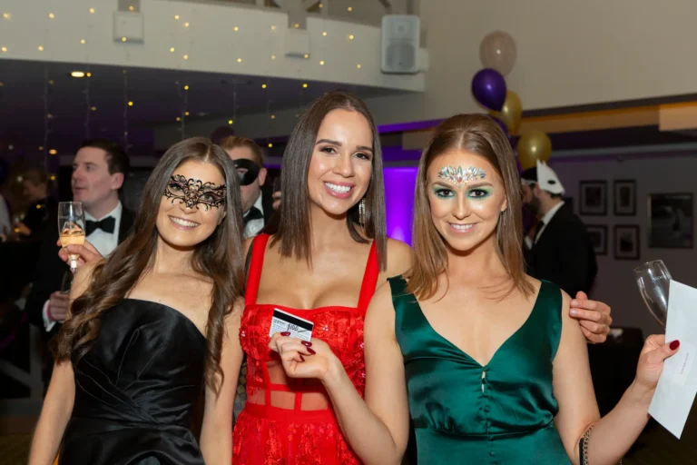 kooyong lawn tennis club masquerade ball guest award moment Three women pose together during the Kooyong Lawn Tennis Club End-of-Year Masquerade Ball, dressed in elegant evening wear and masquerade-style masks or decorative face paint. The woman on the left wears a black strapless dress with a black lace mask, the center woman wears a red dress with a matching lace mask and gold belt, holding a small white card, and the woman on the right wears a green V-neck dress with intricate white face paint. They stand in front of a white and cream artificial rose wall, with purple lighting, balloon clusters, and mingling guests visible in the background, capturing a festive and celebratory moment during the speeches.