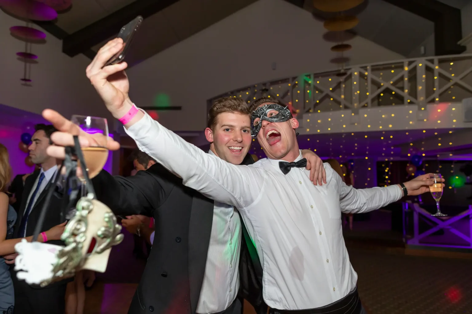 Two men pose joyfully at the Kooyong Lawn Tennis Club End-of-Year Masquerade Ball in Toorak. The man on the left, in a black tuxedo jacket over a white shirt, holds up his phone to take a selfie while clutching two masquerade masks and wearing a pink event wristband. The man on the right, wearing a tuxedo shirt, black bow tie, and silver-and-black masquerade mask, spreads his arms wide in celebration, holding a glass of champagne and smiling broadly. The indoor venue features a mezzanine with white crisscross railings, strings of warm fairy lights, purple and gold balloons, and guests in formal attire mingling in the background.