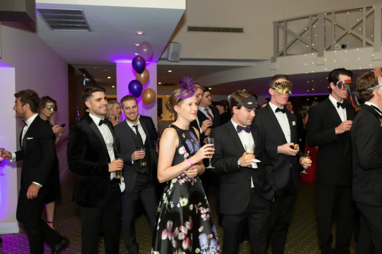 kooyong lawn tennis club masquerade ball guests standing during speech Guests stand together inside the Kooyong Lawn Tennis Club clubhouse during a formal speech at the End-of-Year Masquerade Ball, dressed in elegant black-tie attire and masquerade masks while holding drinks, surrounded by purple and gold balloon décor, glass partitions, and classic interior details, capturing an attentive, social moment during this prestigious and celebratory Melbourne event.