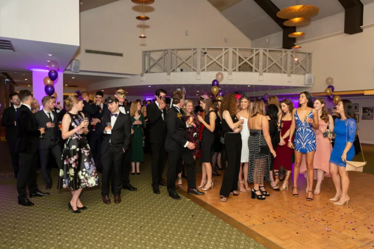 kooyong lawn tennis club masquerade ball guests standing during speeches Guests gather standing in the main event space during the formal speeches at the Kooyong Lawn Tennis Club End-of-Year Masquerade Ball, dressed in elegant black-tie attire and surrounded by rose wall décor, balloon installations, and the club’s grand interior, capturing a shared moment of attention, celebration, and refined atmosphere at this prestigious end-of-year event in Melbourne.