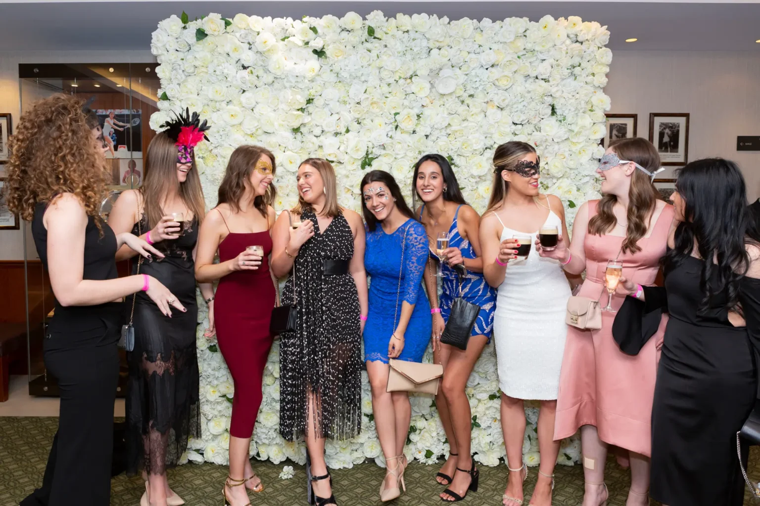 Nine women pose together at the YarraYong Ball at Kooyong Lawn Tennis Club in Toorak, smiling in front of a floor-to-ceiling white rose floral wall. Dressed in elegant black-tie gowns in black, red, blue, pink and white, and wearing colourful masquerade masks, they hold drinks and enjoy a lively moment inside the prestigious club, with tennis memorabilia and reflective walls adding to the sophisticated event atmosphere.