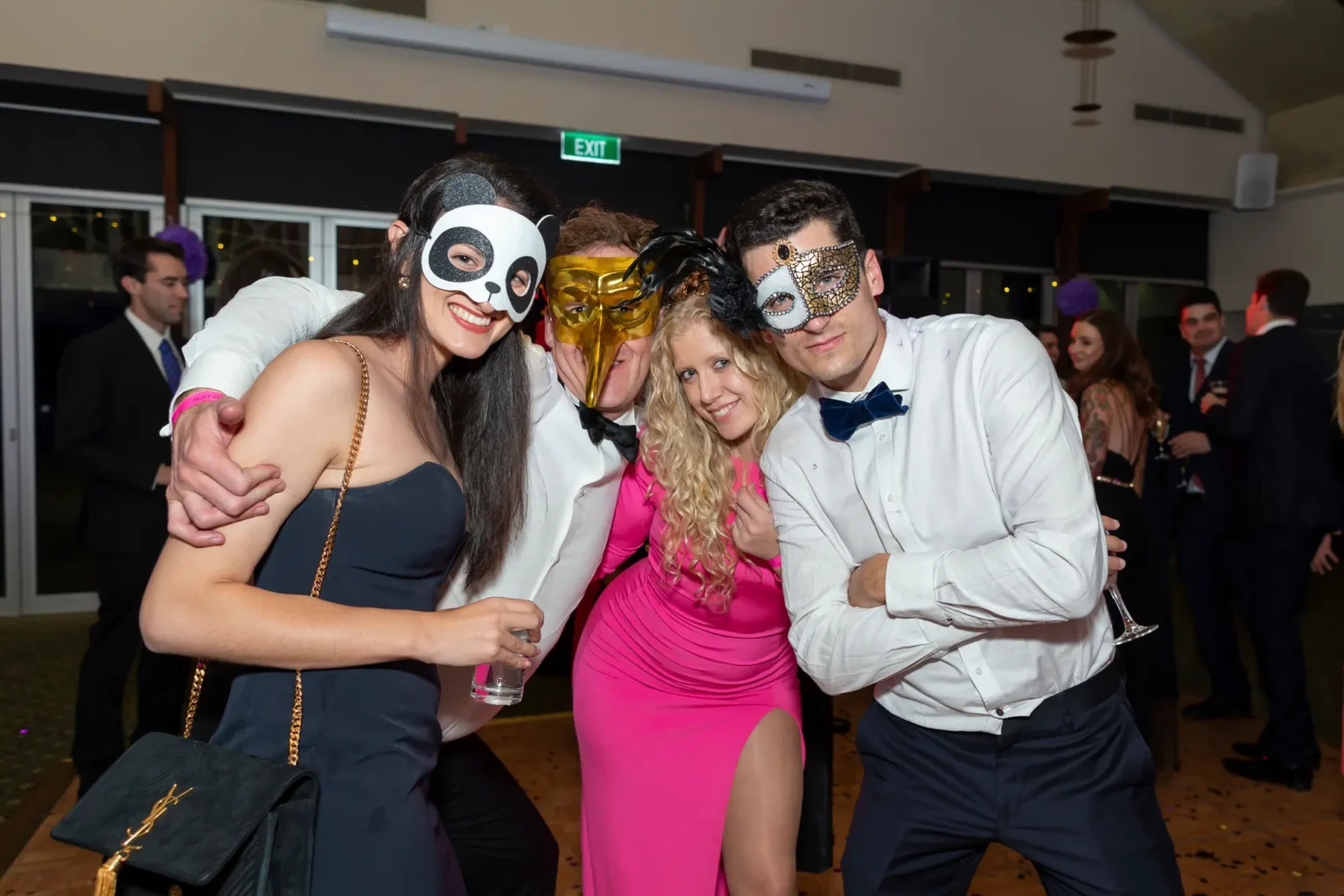Four guests pose together at the Kooyong Lawn Tennis Club End-of-Year Masquerade Ball, featuring a woman in a panda mask, a man in a gold bird-beak mask, a woman in a bright fuchsia pink dress, and a man in a silver mask. The dark wood–paneled function room and mingling guests create a lively black-tie party atmosphere.