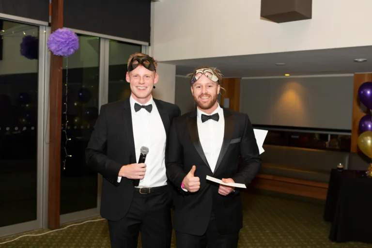 kooyong lawn tennis club masquerade ball prize presentation Two men at the Kooyong Lawn Tennis Club End-of-Year Masquerade Ball in Melbourne celebrate during a prize presentation. The man on the left, dressed in a black tuxedo with a black bow tie and masquerade eye mask, holds a microphone and smiles at the camera, while the man on the right gives a thumbs-up gesture, holding a white card. The indoor venue features a dark patterned carpet over a red runner, glass partitions, purple pom-pom decorations, ambient purple lighting, balloon clusters, and professional audio equipment, capturing a festive, elegant, and social moment at the formal masquerade event.