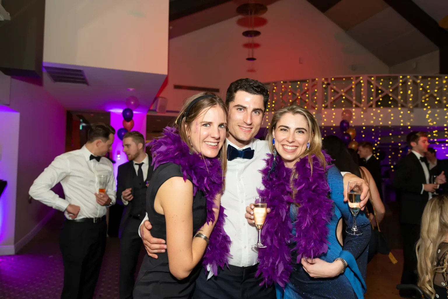 Three guests pose closely together wearing purple feather boas at the Kooyong Lawn Tennis Club End-of-Year Masquerade Ball, smiling and holding champagne as they enjoy the lively celebration. Surrounded by fairy lights, purple and gold balloons, and elegantly dressed guests, the image captures the energetic and social atmosphere of the black-tie year-end event.