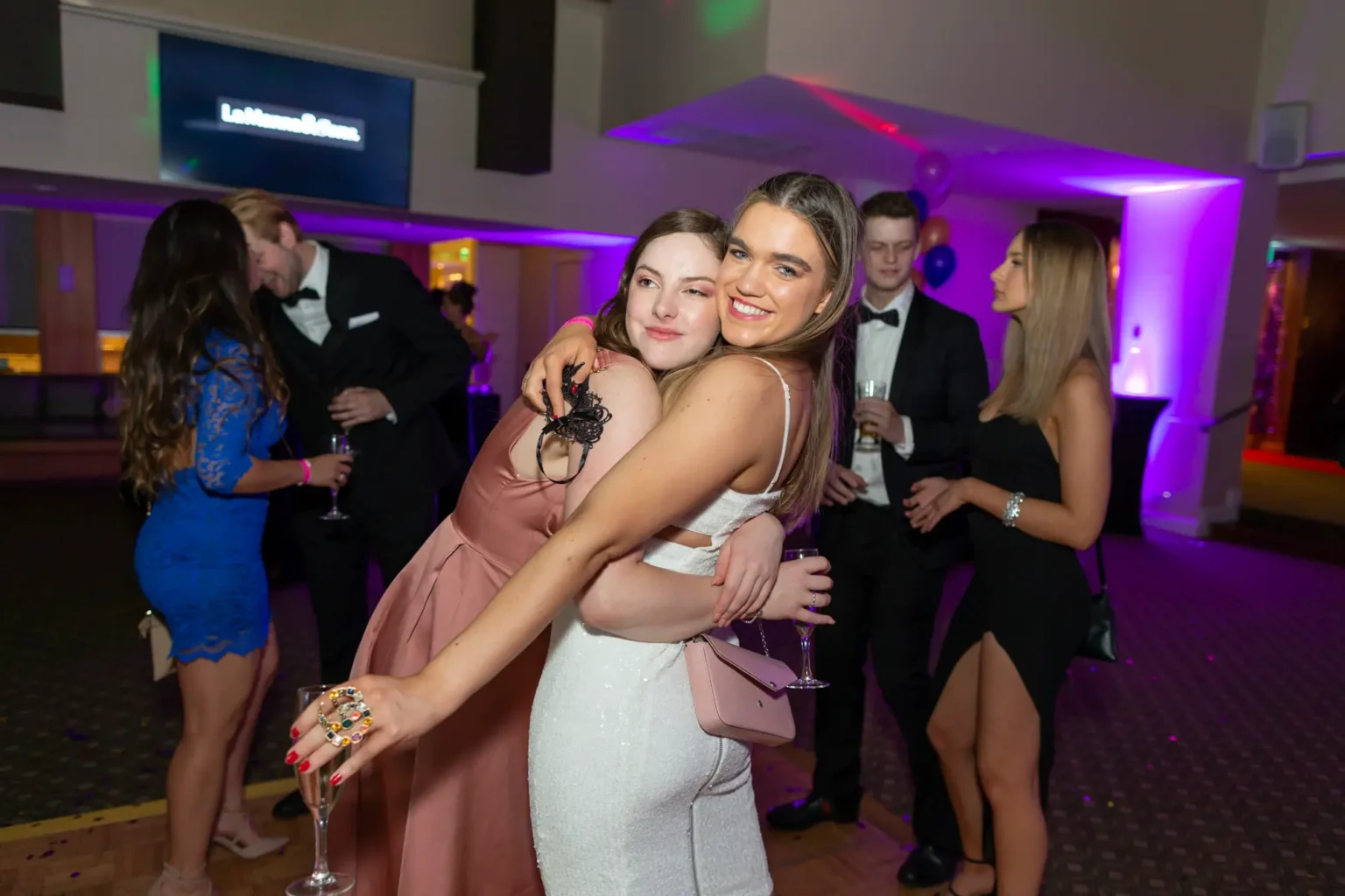 Four guests enjoy the Kooyong Lawn Tennis Club End-of-Year Masquerade Ball in Toorak. A woman in a royal blue lace dress holds a glass on the left, while two women in the center embrace—one in a rose-pink dress, the other in a white sequined dress with spaghetti straps. On the right, a woman in a black mini-dress with a thigh-high slit smiles with a glass in hand. A man in a black tuxedo stands slightly behind, smiling. The indoor function room features purple accent lighting, patterned carpet, balloons, and mingling guests, creating a festive and joyful atmosphere.