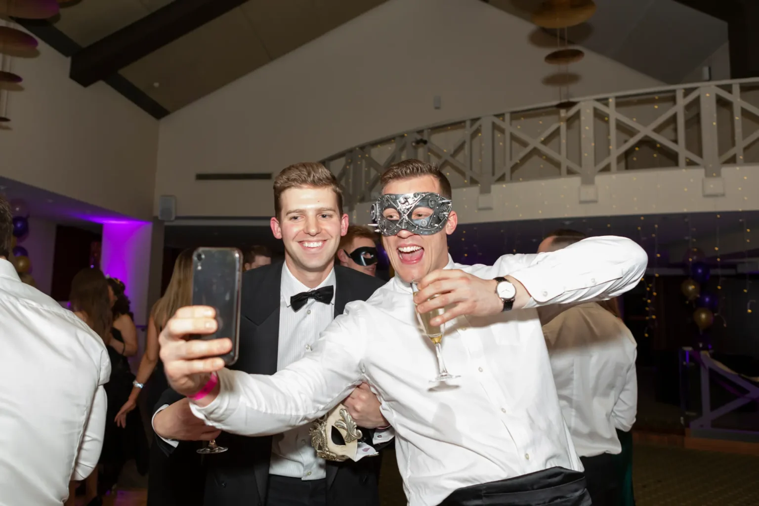Two men take a lively selfie at the Kooyong Lawn Tennis Club End-of-Year Masquerade Ball, one wearing a silver masquerade mask with arms outstretched holding champagne, while the other smiles in a tuxedo. Fairy lights, purple and gold balloons, and mingling guests fill the background, capturing the energetic and social atmosphere of the black-tie celebration.