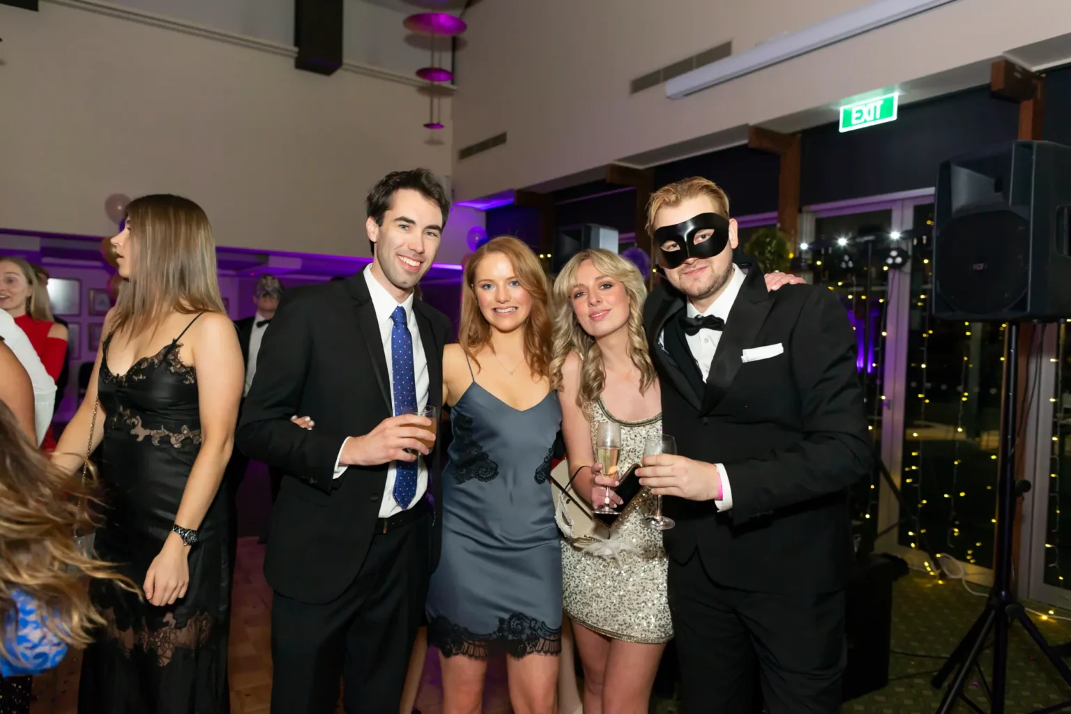 Four guests pose together at the Kooyong Lawn Tennis Club End-of-Year Masquerade Ball, featuring a man in a tuxedo with a blue tie, a woman in a grey-blue lace-trim slip dress, a blonde woman in a sequined mini dress, and a man in a black masquerade mask holding champagne. The elegant wood-paneled function room and mingling guests create a joyful black-tie party atmosphere.