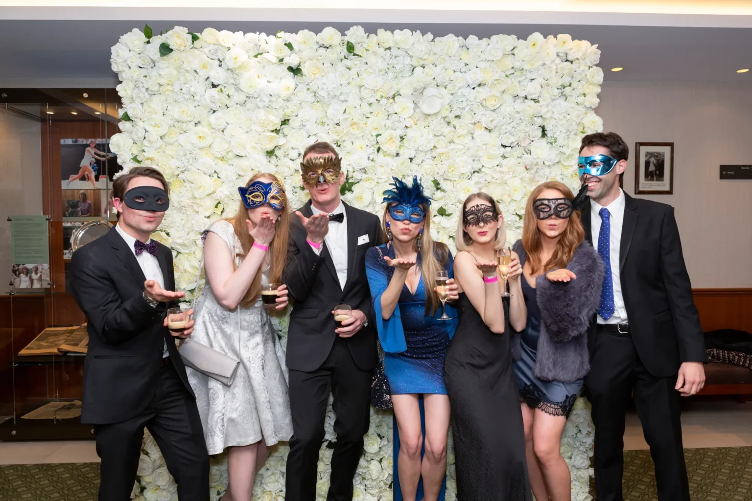 Seven guests pose in front of a white rose floral wall at the YarraYong Ball at Kooyong Lawn Tennis Club in Toorak, smiling and blowing kisses toward the camera. Dressed in elegant black-tie attire in black, silver and blue, and wearing colourful masquerade masks, they hold drinks and enjoy a joyful group moment inside the prestigious clubhouse, with tennis memorabilia and reflective walls enhancing the sophisticated event atmosphere.