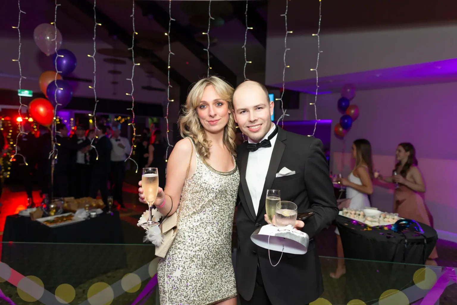 A man and woman pose together at the Kooyong Lawn Tennis Club End-of-Year Masquerade Ball in Toorak. The woman wears a silver sequined dress and holds a champagne flute with a light-colored clutch under her arm. The man wears a black tuxedo with bow tie and pocket square, holding a champagne flute and a tumbler. Strings of fairy lights, colorful balloons, buffet tables, and other guests in formal attire are visible in the blurred indoor background, creating a festive and celebratory atmosphere.