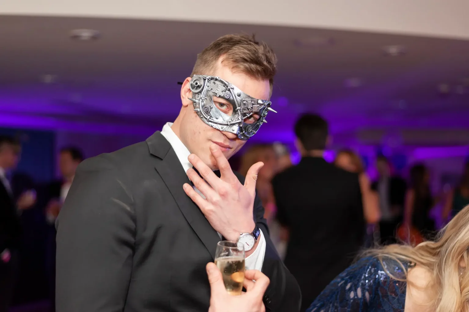 A man poses for a candid portrait at the YarraYong Ball at Kooyong Lawn Tennis Club in Toorak, wearing a black suit and an intricate silver steampunk-style masquerade mask. Purple uplighting and warm ambient lighting create a festive atmosphere, while other guests in black-tie attire mingle in the softly blurred background of the elegant indoor function space.
