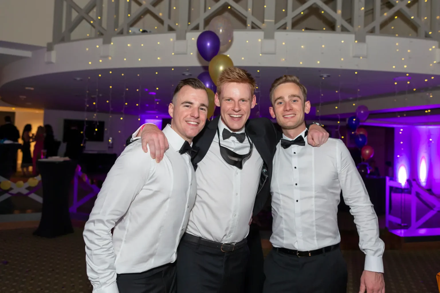 Three men pose together at the Kooyong Lawn Tennis Club End-of-Year Masquerade Ball in Toorak. The man on the left wears a white dress shirt and black trousers, smiling with his arm around the center man. The center man wears a tuxedo jacket, white shirt, black bow tie, and has a black masquerade mask around his neck, smiling broadly. The man on the right is in a white dress shirt, black trousers, and black bow tie, smiling warmly with his arm around the center man's shoulder. The indoor venue features a mezzanine with white crisscross railings, strings of warm fairy lights, clusters of purple, gold, and white balloons, and guests mingling in the background.