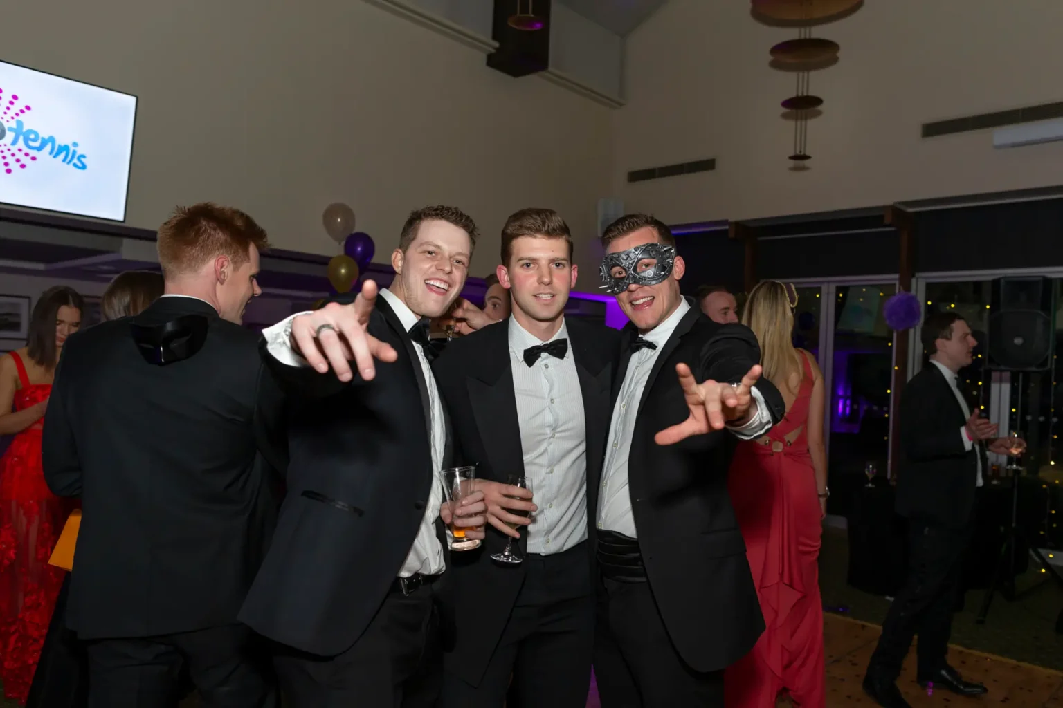 Three men pose energetically at the Kooyong Lawn Tennis Club End-of-Year Masquerade Ball in Toorak. The man on the left crouches slightly, pointing at the camera with a light-colored drink in hand, smiling widely. The center man stands upright in a tuxedo holding champagne. The man on the right wears a silver masquerade mask and mirrors the left man's pose. Guests in formal attire and red dresses mingle in the background, with purple and gold balloons, patterned carpet, and ambient lighting creating a festive indoor atmosphere.