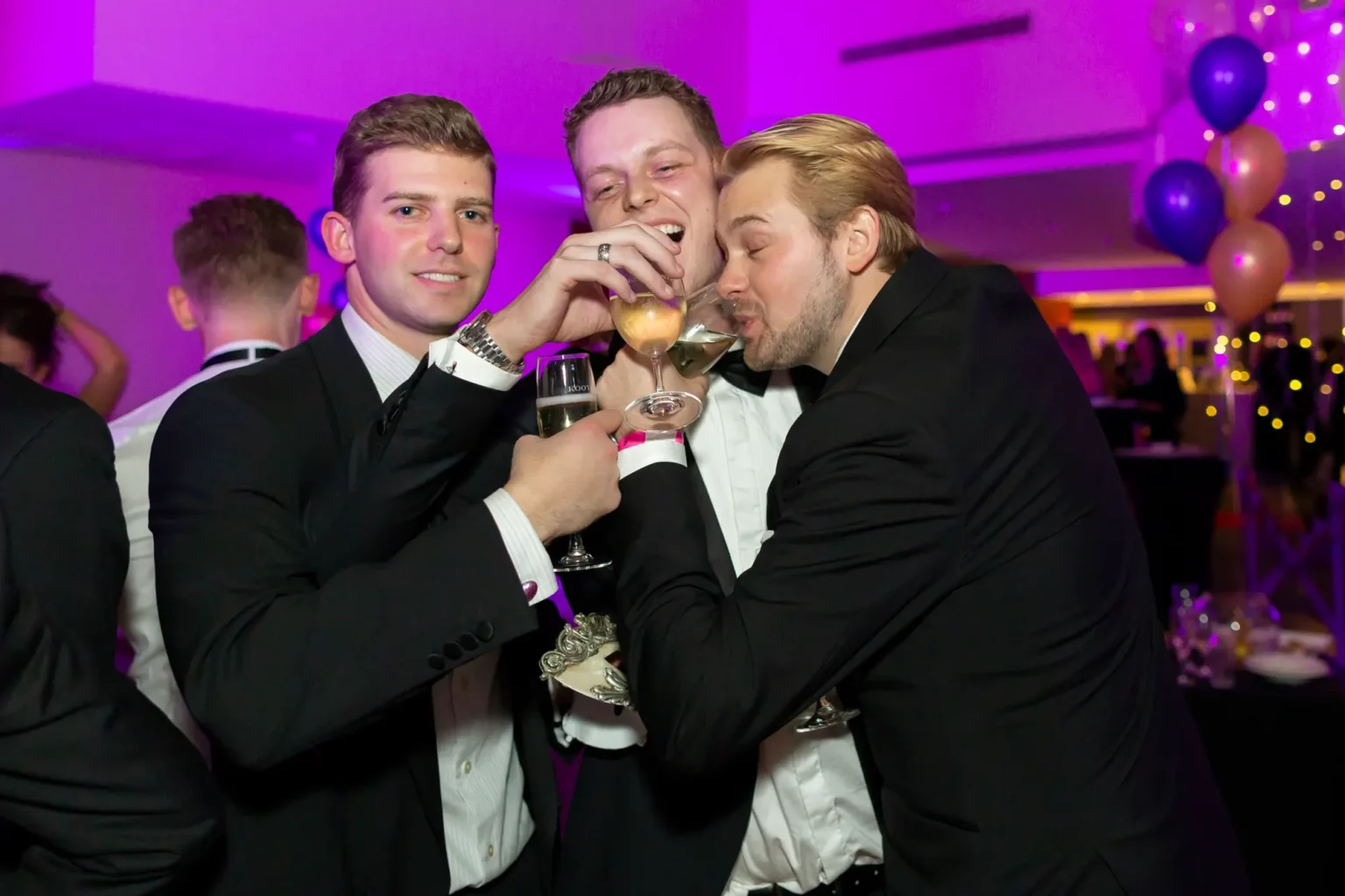 Three men are captured in a lively, celebratory moment at the Kooyong Lawn Tennis Club End-of-Year Masquerade Ball in Toorak. All three wear tuxedos and bow ties. The man on the left smiles warmly at the camera holding a champagne flute, the center man laughs enthusiastically holding a light-colored drink with a pink wristband, and the man on the right leans in drinking from his champagne flute. The indoor venue features purple ambient lighting, clusters of purple and gold balloons, and formally dressed guests mingling in the background, creating a vibrant, festive atmosphere.