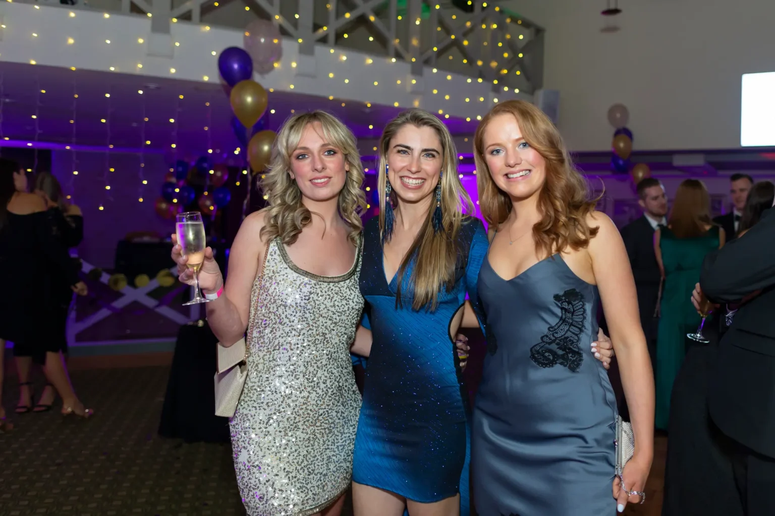 Three women dressed in elegant evening attire pose together at the Kooyong Lawn Tennis Club End-of-Year Masquerade Ball in Toorak. The woman on the left wears a silver sequined dress, the center in a vibrant blue gown, and the right in a grey dress with black lace. Fairy lights, balloons, and mingling guests create a festive, joyful atmosphere.