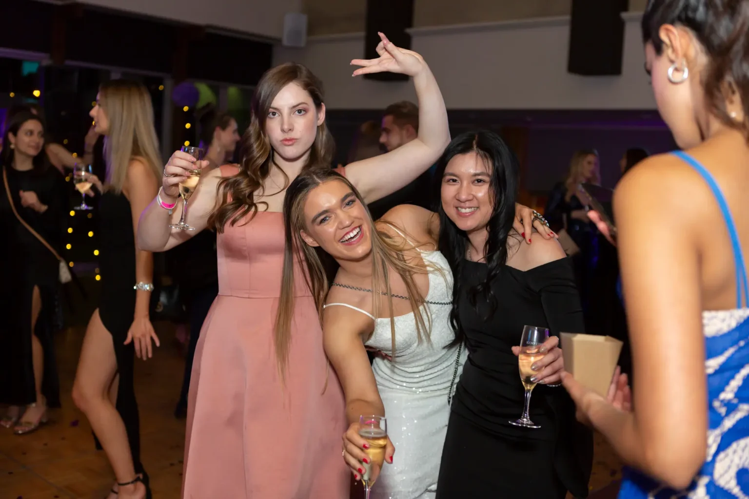 Three women pose joyfully together at the Kooyong Lawn Tennis Club End-of-Year Masquerade Ball, smiling and holding champagne flutes. One woman in a pink dress raises her arm playfully, the center woman in a white sequined dress leans forward laughing, and the third woman in an off-the-shoulder black dress smiles warmly with her arm around her friend. The indoor evening venue features warm ambient lighting, a dark wooden floor, and other formally dressed guests mingling in the background, creating a glamorous and celebratory atmosphere.