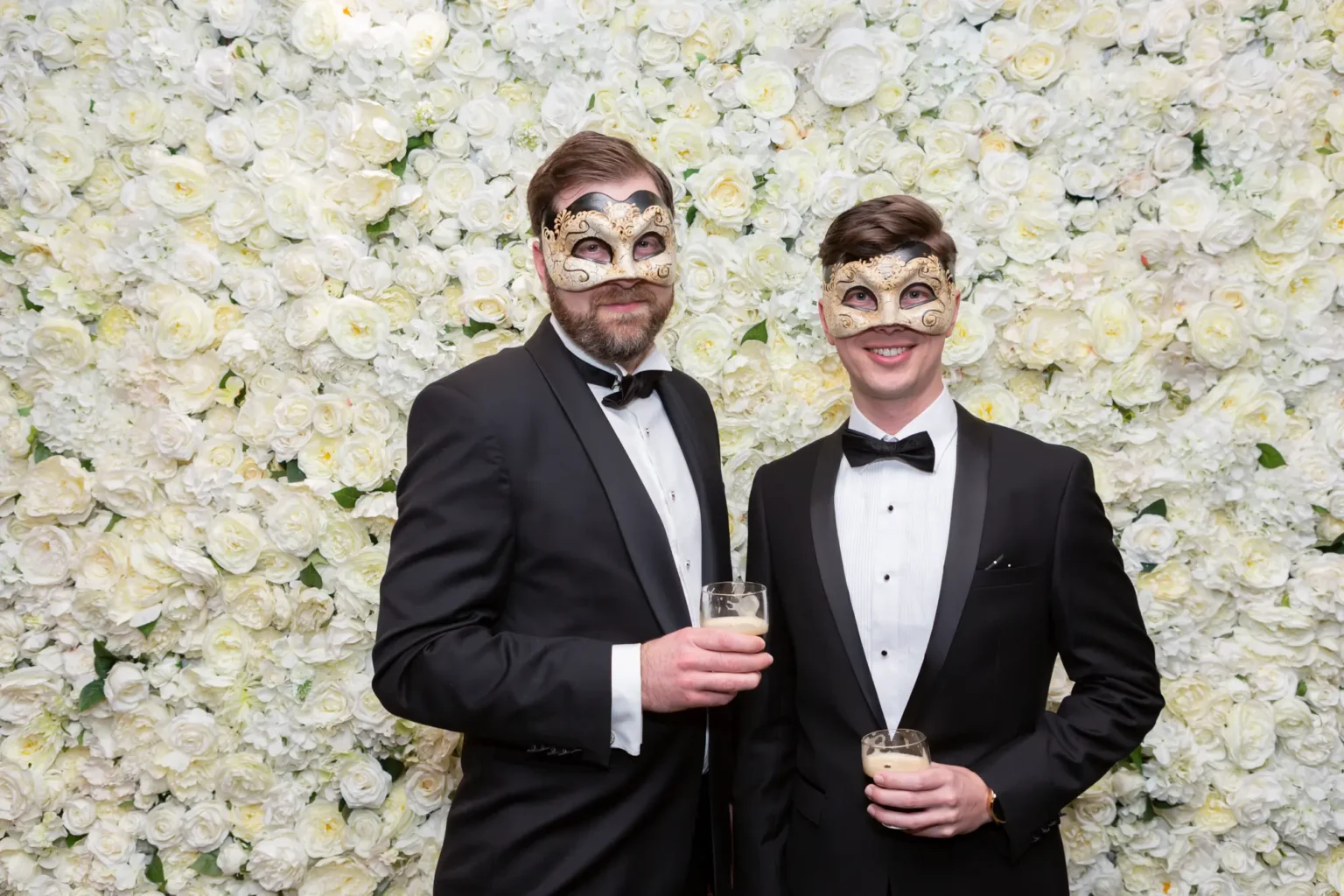 Two men pose together in front of a white rose floral wall at the YarraYong Ball at Kooyong Lawn Tennis Club in Toorak, smiling and holding champagne glasses. Dressed in black tuxedos with gold and black masquerade masks, they share a stylish and joyful moment during the elegant end-of-year celebration inside the prestigious clubhouse.