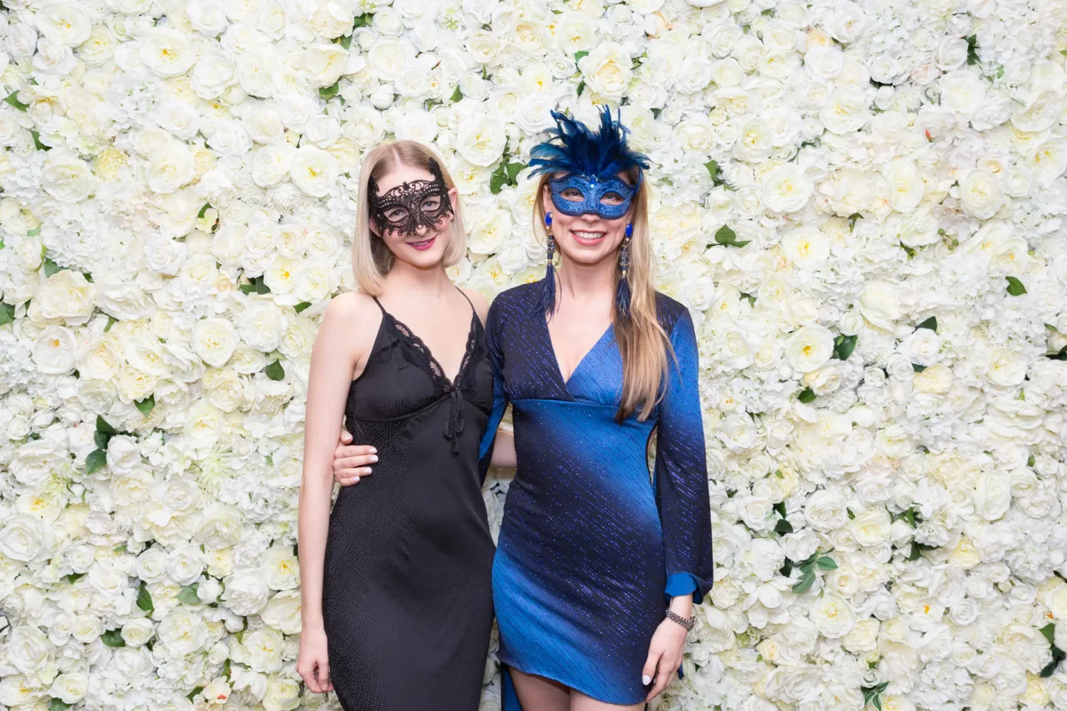 Two women pose together at the YarraYong Ball at Kooyong Lawn Tennis Club in Toorak, smiling in front of a white rose floral wall. One wears a black dress with a lace mask while the other wears a blue sequined gown with a vibrant blue feathered masquerade mask, holding a drink in the elegant, warmly lit indoor function space.