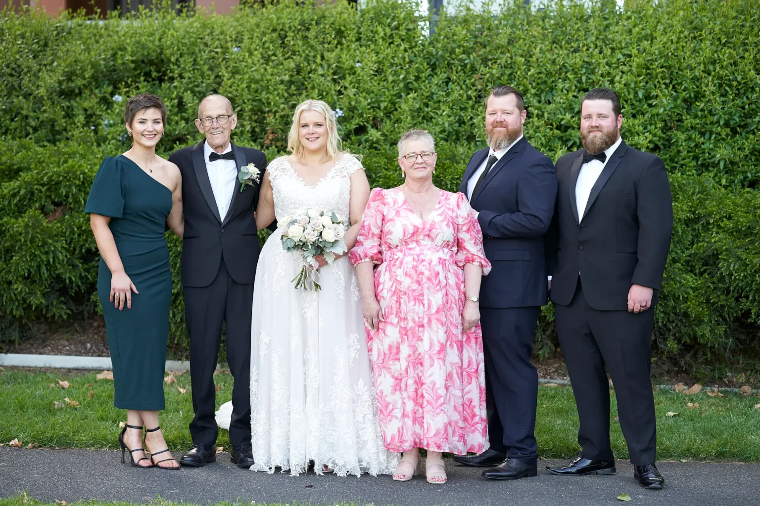 Bride stands with her parents and siblings during a formal family portrait in Burnley Park near Amora Herencia Riverwalk Melbourne. The bride in a lace A-line wedding gown holds a bouquet of blush and white roses, while her father wears a black tuxedo, her mother a pink floral dress, her sister a forest-green one-shoulder dress, and her brothers wear a navy suit and black tuxedo. A manicured hedge creates a natural Melbourne parkland backdrop.