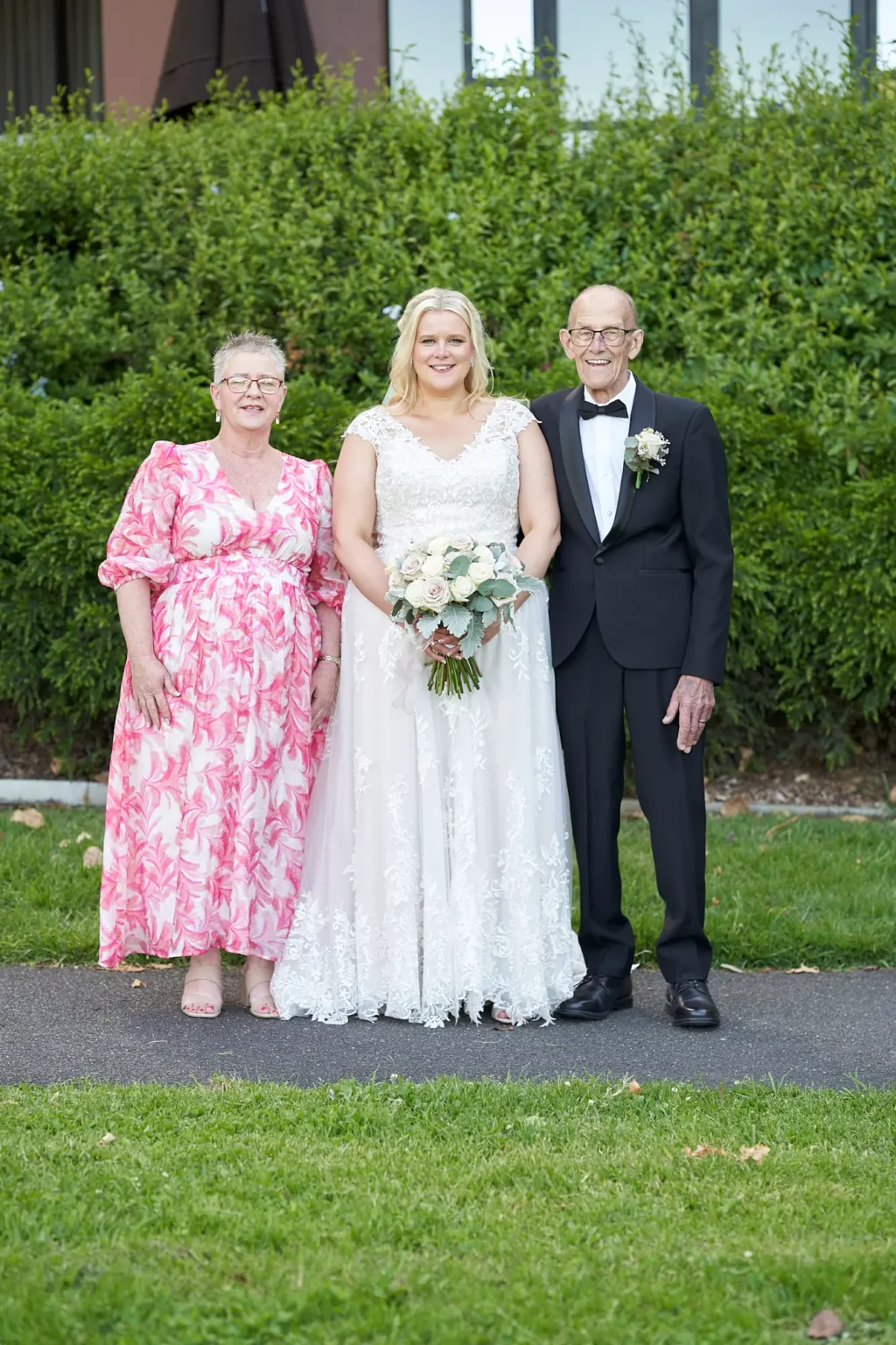 Bride stands between her mother and father during a formal family portrait in Burnley Park near Amora Herencia Riverwalk Melbourne. The bride wears a lace A-line wedding gown and holds a bouquet of white and blush roses, while the father stands in a black tuxedo with a white rose boutonniere and the mother wears a pink floral dress. A manicured green hedge and the Amora Riverwalk hotel building create an elegant Melbourne parkland backdrop.