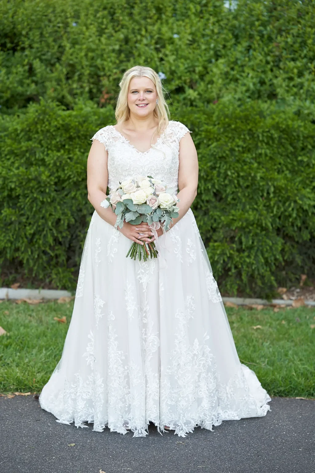 Bride stands for an elegant bridal portrait in Burnley Park near Amora Herencia Riverwalk Melbourne. The bride wears a lace A-line wedding gown with floral appliqué and holds a bouquet of white and blush roses with eucalyptus. She smiles toward the camera while standing on a garden path with a manicured green hedge creating a bright and classic Melbourne parkland wedding backdrop.