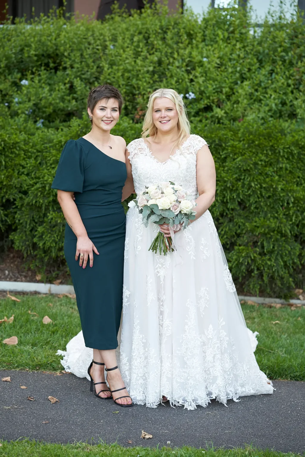Bride stands with her sister during a formal wedding portrait in Burnley Park near Amora Herencia Riverwalk Melbourne. The bride wears a lace A-line wedding gown and holds a bouquet of blush and white roses, while her sister wears a forest-green one-shoulder midi dress with black strappy heels. The pair stand closely together on a garden path with a manicured green hedge creating an elegant Melbourne parkland backdrop.