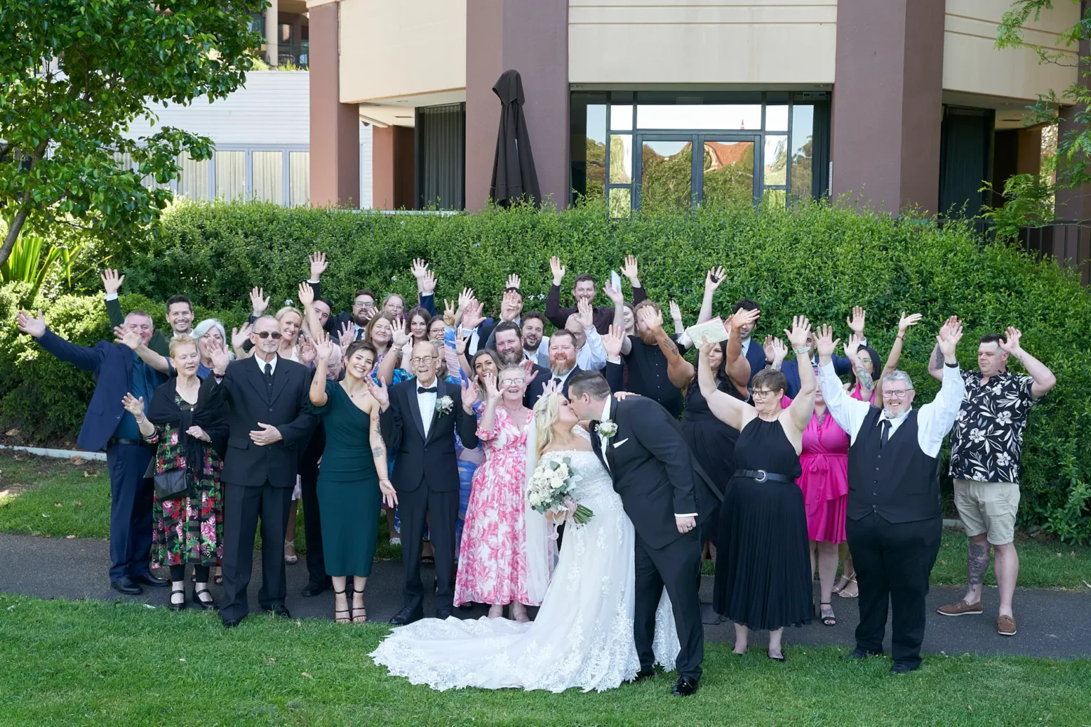 Bride and groom kiss at the center of a lively large group wedding portrait in Burnley Park near Amora Herencia Riverwalk Melbourne. The bride in a lace A-line wedding gown holds a bouquet while the groom in a black tuxedo embraces her. Family and friends stand around the couple with raised arms and joyful cheers, framed by a manicured green hedge and the modern Amora Riverwalk hotel building in Richmond.