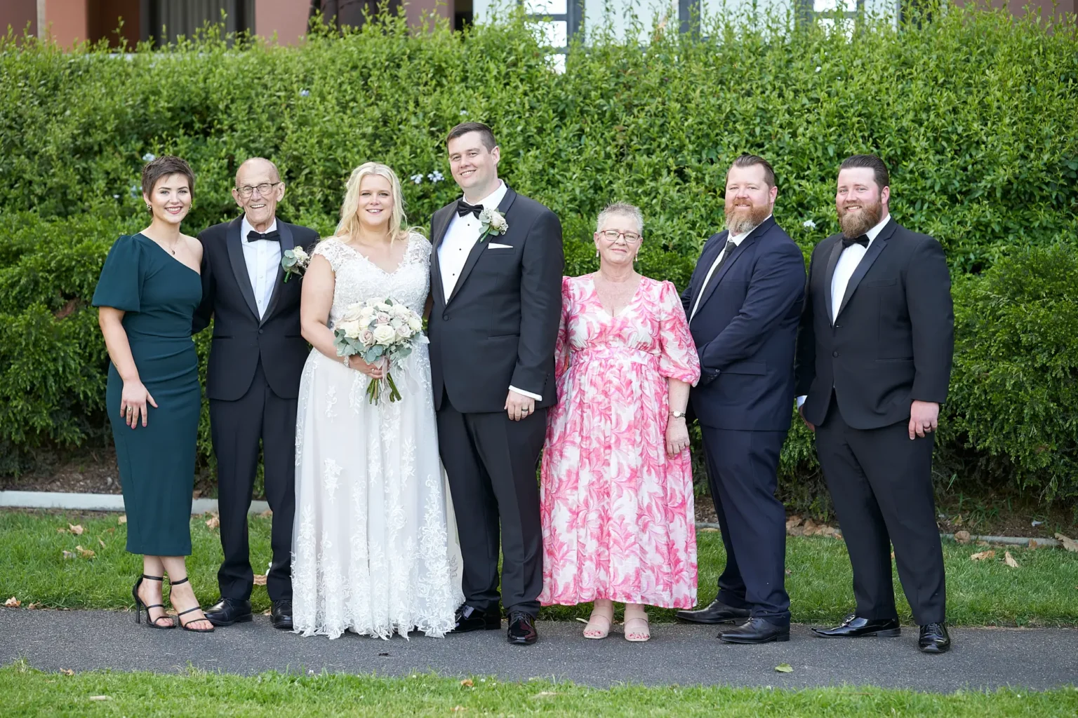 Bride and groom stand with the bride’s parents and siblings during a formal family portrait in Burnley Park near Amora Herencia Riverwalk Melbourne. The bride wears a lace A-line wedding gown and holds a bouquet of blush and white roses while the groom stands in a black tuxedo. The bride’s mother wears a pink floral dress, her father a black tuxedo, her sister a forest-green one-shoulder dress, and her brothers wear a navy suit and black tuxedo, framed by a manicured hedge in the Melbourne parkland setting.