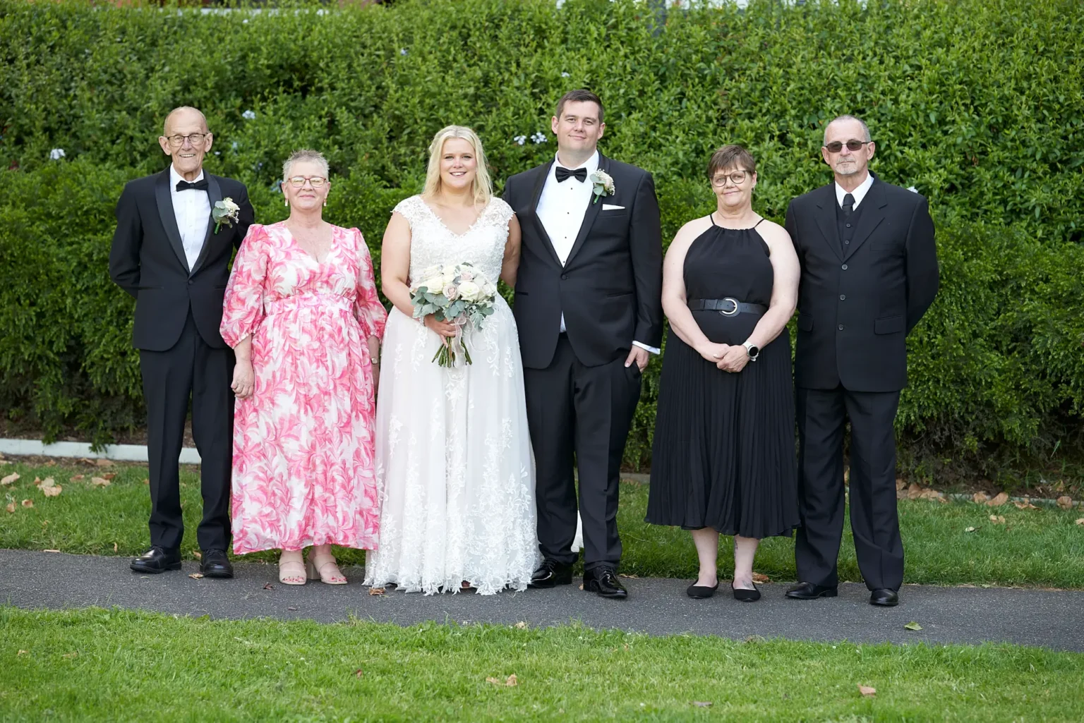 Bride and groom stand with both sets of parents during a formal family portrait in Burnley Park near Amora Herencia Riverwalk Melbourne. The bride wears a lace A-line wedding gown and holds a bouquet of blush and white roses while the groom stands in a black tuxedo with a white rose boutonniere. The bride’s parents and groom’s parents stand beside them in elegant formal attire, framed by a manicured green hedge in the Melbourne parkland setting.