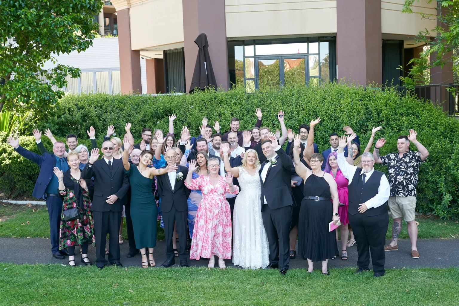 Bride and groom celebrate with a large group of family and friends during a joyful wedding group portrait in Burnley Park near Amora Herencia Riverwalk Melbourne. The bride in a lace wedding gown and the groom in a black tuxedo stand at the center while guests raise their arms and cheer toward the camera. A manicured green hedge and the modern Amora Riverwalk hotel building create a vibrant Melbourne parkland backdrop.