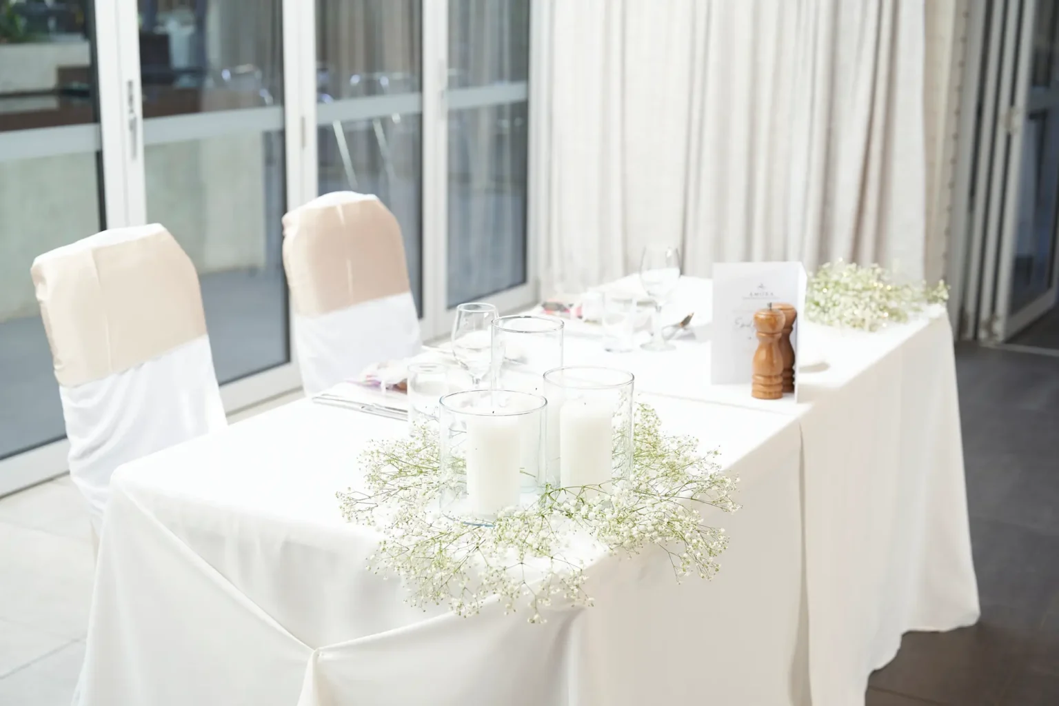 Bride and groom sweetheart table at Amora Herencia Riverwalk Melbourne wedding reception featuring a white linen table with baby’s breath floral centerpiece, pillar candles in glass cylinders, menu stationery, and champagne-gold chair sashes, set beside large glass doors overlooking the courtyard.