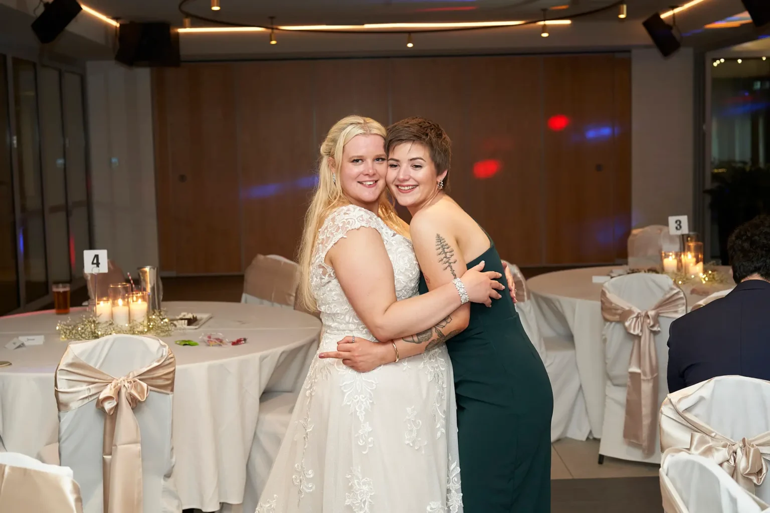 Bride shares a warm embrace with her sister on the dance floor during the wedding reception at Amora Herencia Riverwalk Melbourne. Smiling brightly toward the camera, the bride in her lace A-line wedding gown and her sister in a green dress celebrate together beside elegantly decorated reception tables, capturing a heartfelt family moment during the Melbourne wedding celebration.