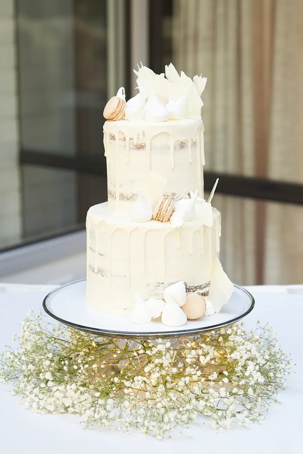 Two-tier semi-naked wedding cake with white chocolate drip, macarons, meringues, and white chocolate shards displayed on a glass stand surrounded by baby’s breath flowers and fairy lights at the Amora Herencia Riverwalk Melbourne wedding reception.