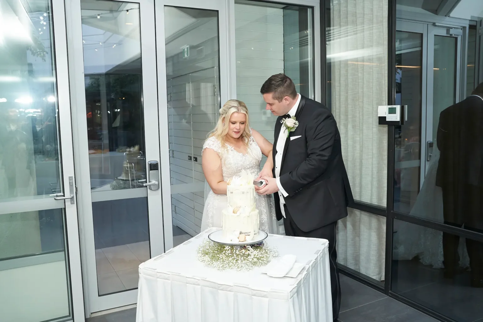 Bride and groom cutting their two-tier white drip wedding cake at Amora Herencia Riverwalk Melbourne reception, standing together with a cake knife beside a baby’s breath floral cake display in the bright Event Pavilion.