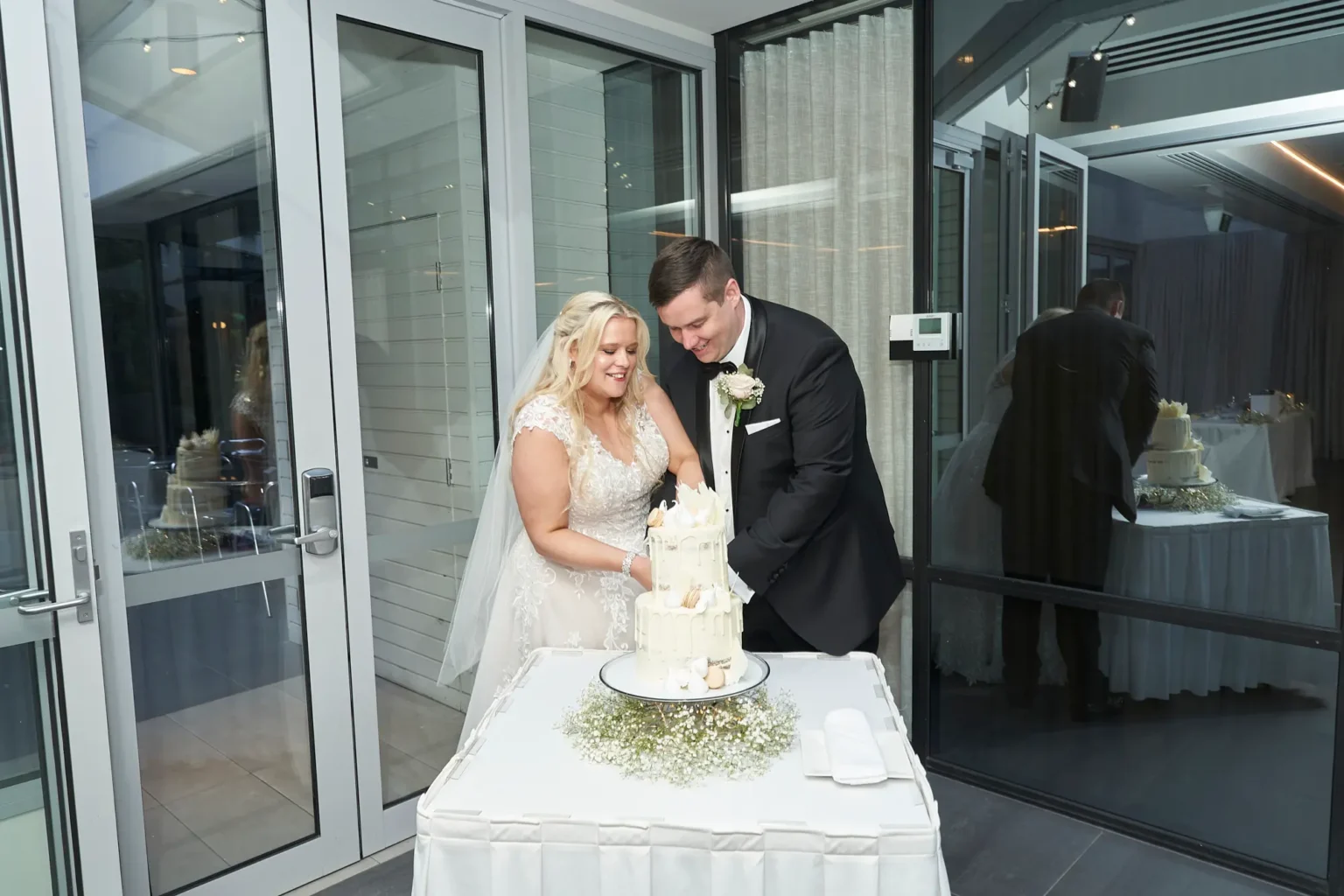 Bride and groom share a meaningful cake cutting moment during their wedding reception at Amora Herencia Riverwalk Melbourne. Standing together in the bright Event Pavilion, they hold the knife hand-in-hand to cut a two-tier white drip wedding cake decorated with macarons and white chocolate shards, displayed on a baby’s breath floral cake table surrounded by soft reception lighting.