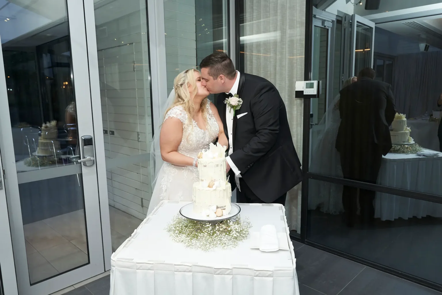 Bride and groom share a romantic kiss beside their wedding cake during the reception at Amora Herencia Riverwalk Melbourne. Standing at the cake table in the bright Event Pavilion, the bride in a lace A-line gown and the groom in a black tuxedo celebrate after cutting their two-tier white drip wedding cake displayed on a baby’s breath floral stand.
