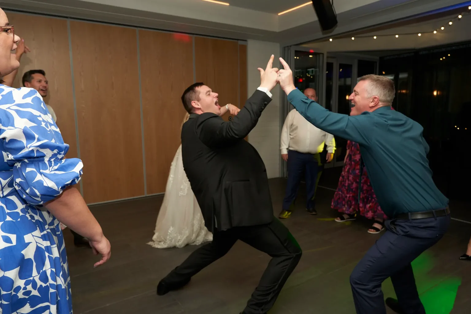 Groom celebrates with a friend on the dance floor during the lively wedding reception at Amora Herencia Riverwalk Melbourne. Wearing a black tuxedo, the groom raises a playful hand gesture while dancing beside his friend in a blue shirt, as other guests and the bride enjoy the high-energy party atmosphere of the Melbourne wedding celebration.