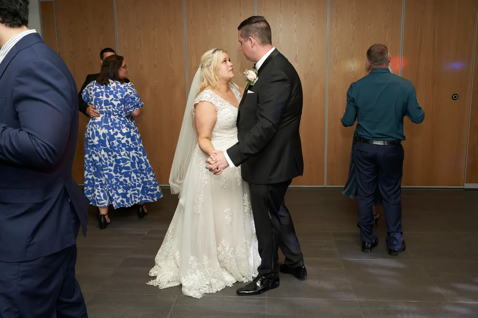 Bride and groom share a romantic slow dance while guests join the dance floor during the wedding reception at Amora Herencia Riverwalk Melbourne. Holding hands and gazing into each other’s eyes, the bride in a lace A-line wedding gown and the groom in a black tuxedo enjoy an intimate moment surrounded by friends and family celebrating their Melbourne wedding.