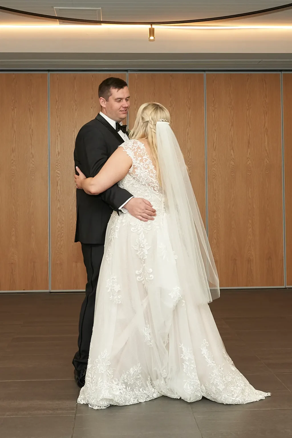 Bride and groom share their romantic first dance during the wedding reception at Amora Herencia Riverwalk Melbourne. The groom holds the bride gently at her waist as they slow dance together, while the bride in a lace A-line gown and veil wraps her arms around his neck, creating an intimate and emotional moment in the softly lit reception space.
