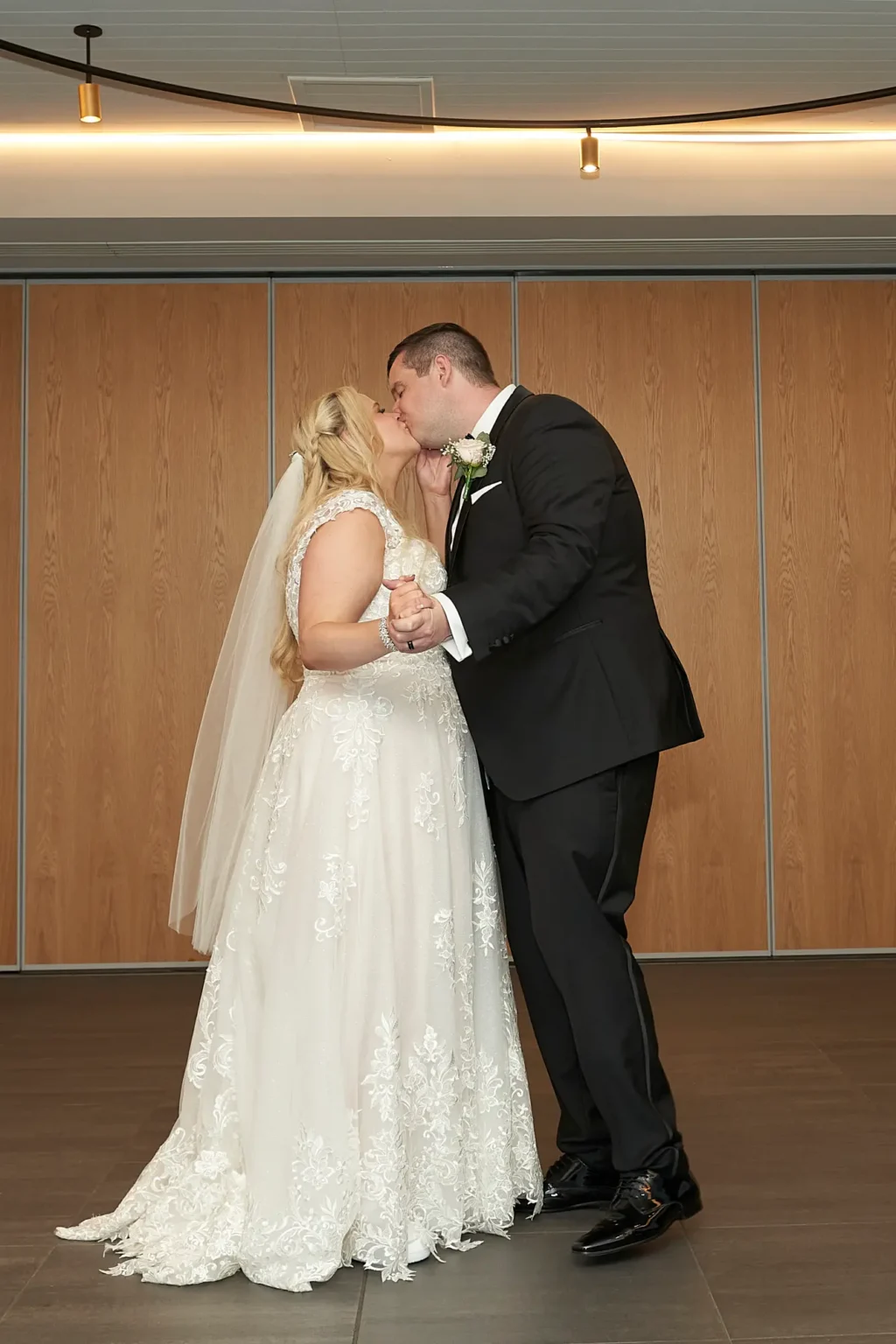 Bride and groom share a romantic kiss during their first dance at the wedding reception at Amora Herencia Riverwalk Melbourne. The groom in a black tuxedo holds the bride’s hands as they dance together, while the bride’s lace A-line gown and train spread elegantly across the floor beneath warm reception lighting, creating an intimate and emotional wedding moment.