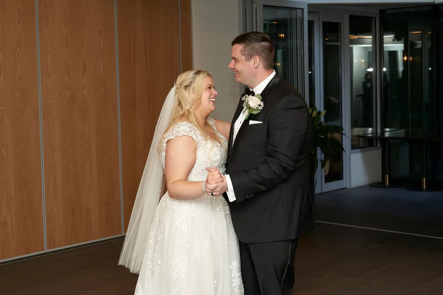Bride and groom laugh together while sharing their first dance at the wedding reception at Amora Herencia Riverwalk Melbourne. Holding hands and smiling into each other’s eyes, the bride in a lace A-line wedding gown and the groom in a black tuxedo enjoy a joyful, intimate moment on the dance floor under warm reception lighting.
