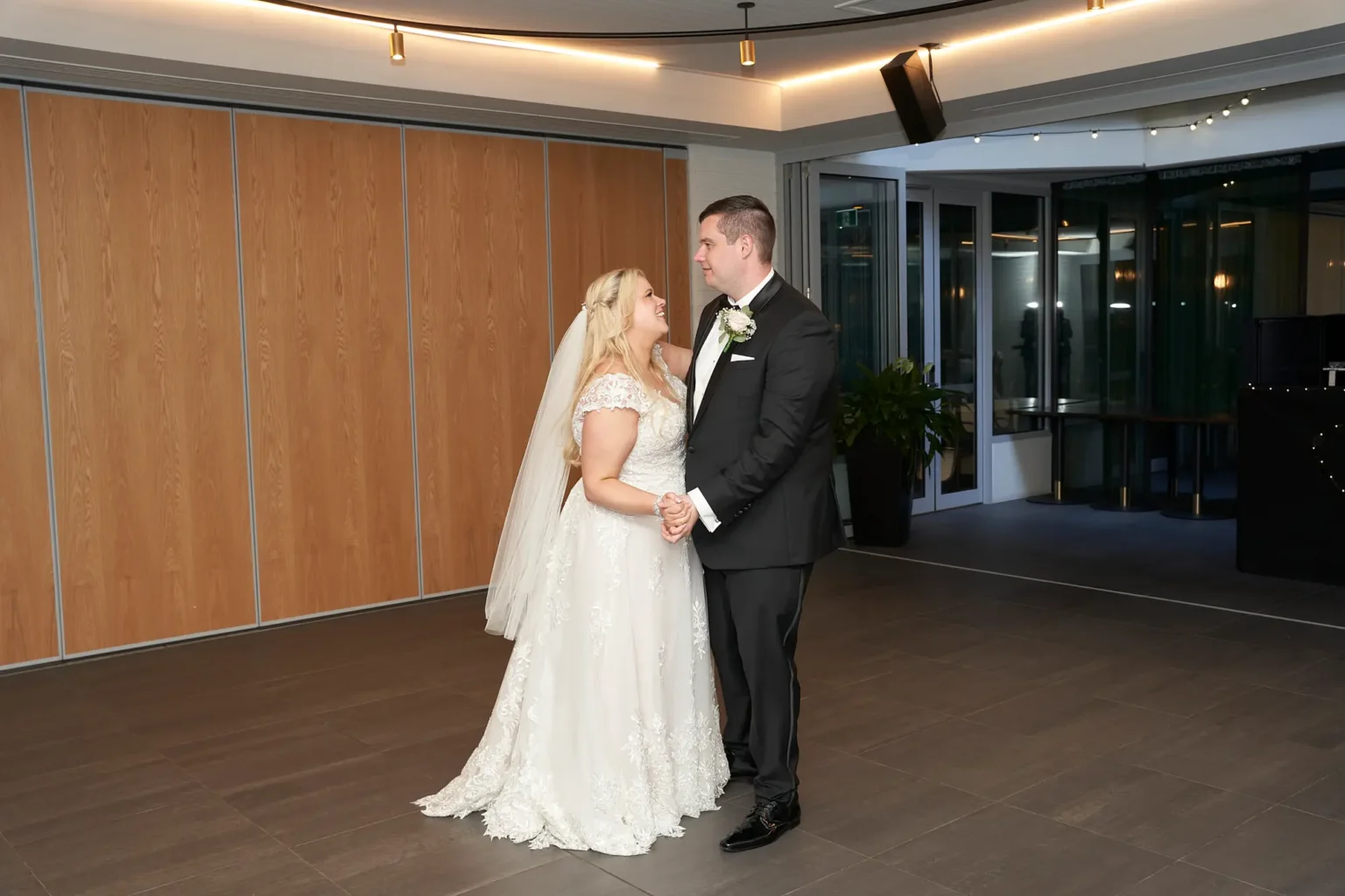 Bride and groom share a romantic first dance at their wedding reception at Amora Herencia Riverwalk Melbourne, holding hands and smiling into each other’s eyes on the dance floor. The bride wears a lace A-line wedding gown with a veil while the groom stands in a black tuxedo, creating an intimate and joyful moment during their elegant Melbourne wedding celebration.